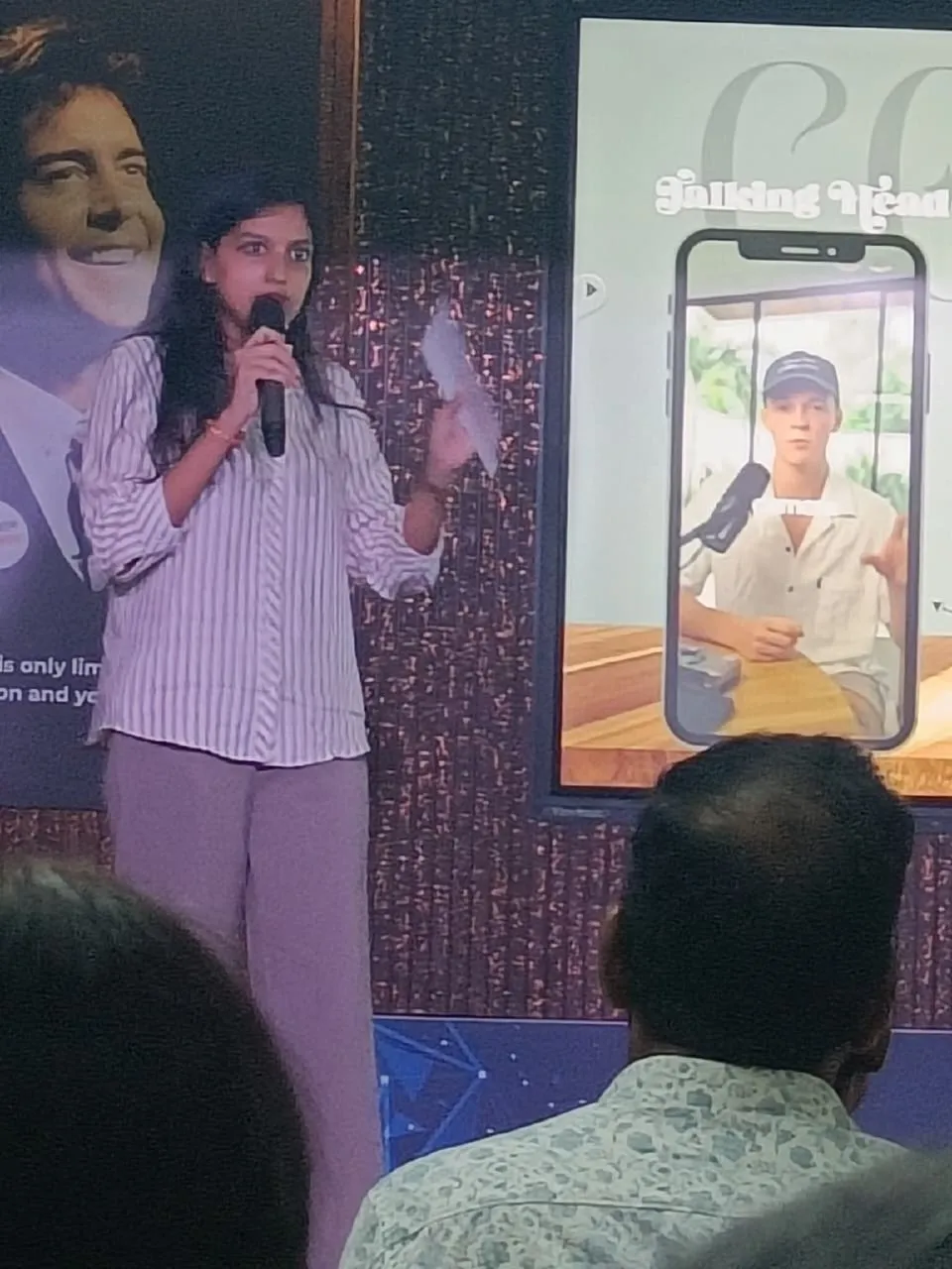 Saijal speaking into a microphone beside a screen showing a talking-head video